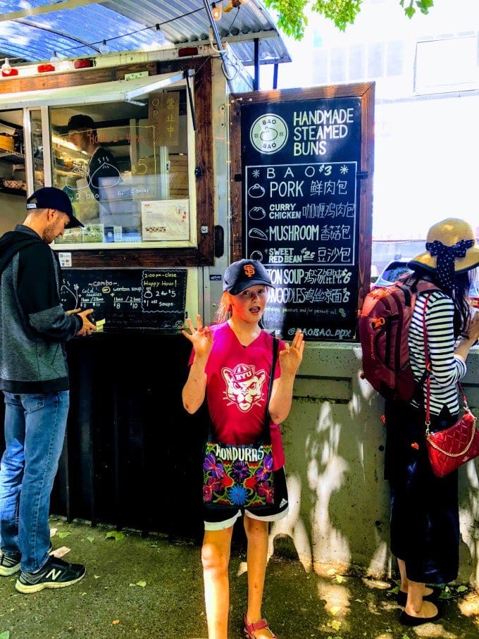 Lucy standing in front of a restaurant sign advertising Handmade Steamed Buns, Bao