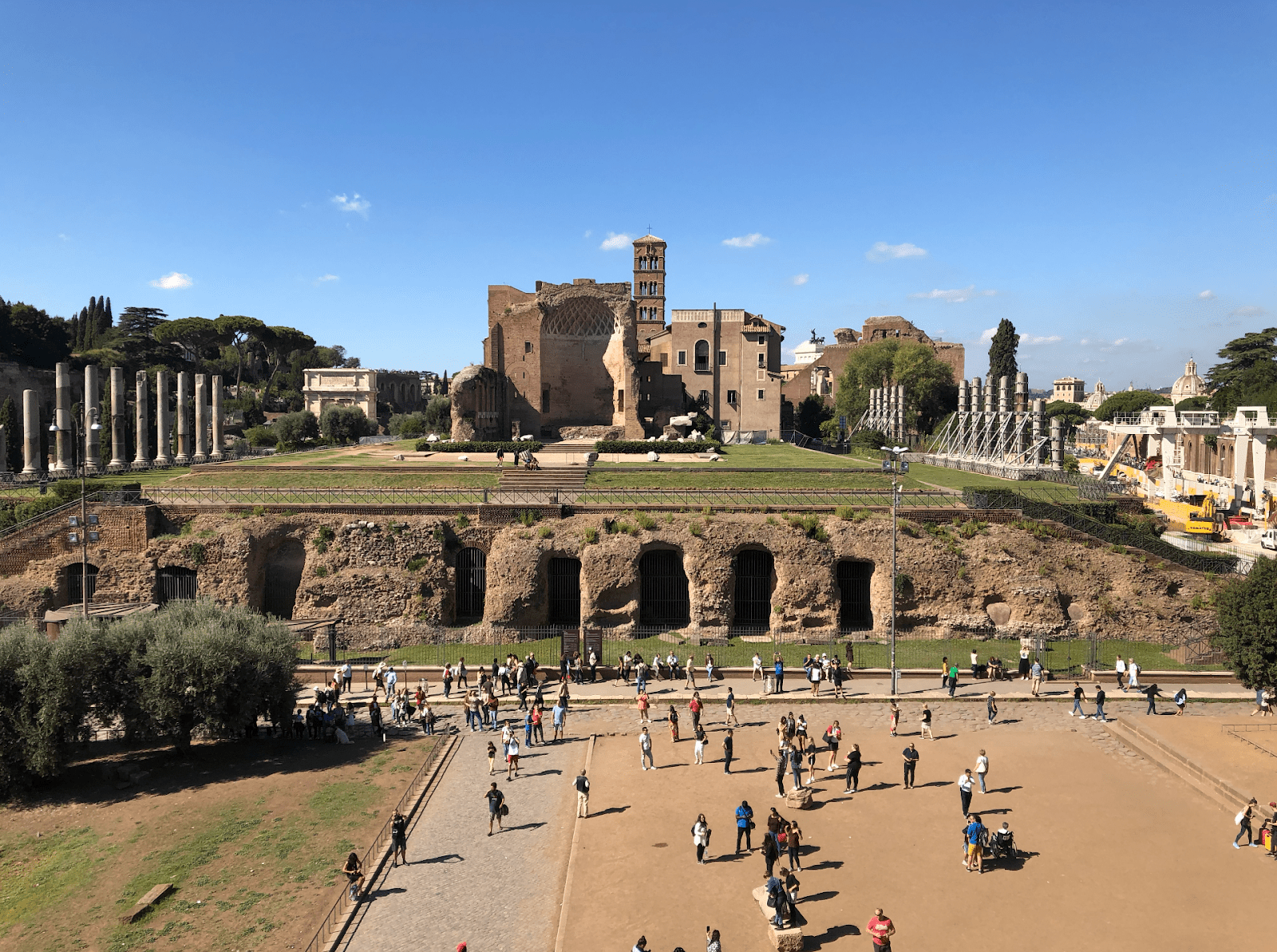 Visitors walk through a broad archaeological area lined with ancient stone ruins in Rome.