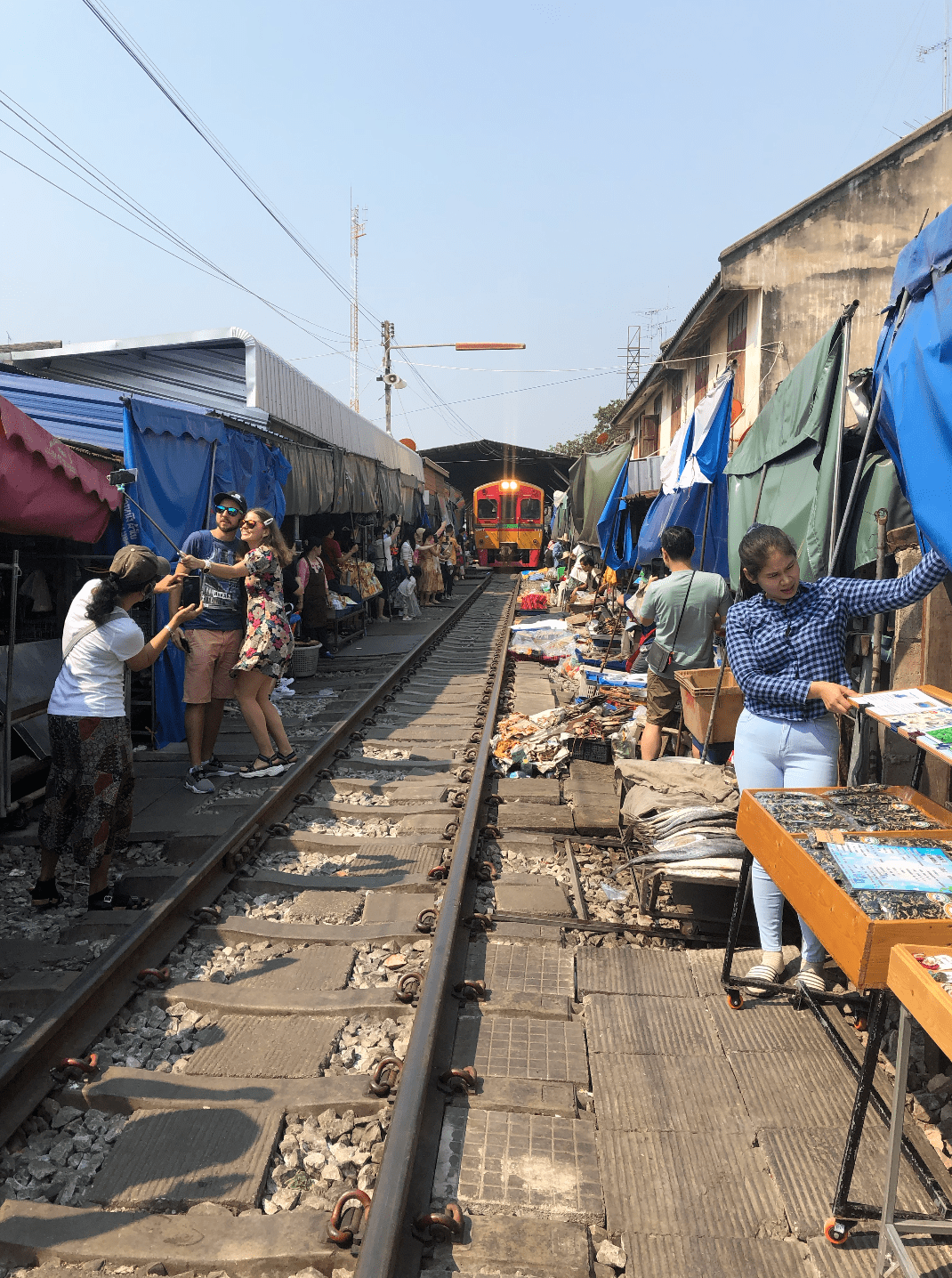 A train approaches through Maeklong market as vendors and visitors stand beside the railway tracks.