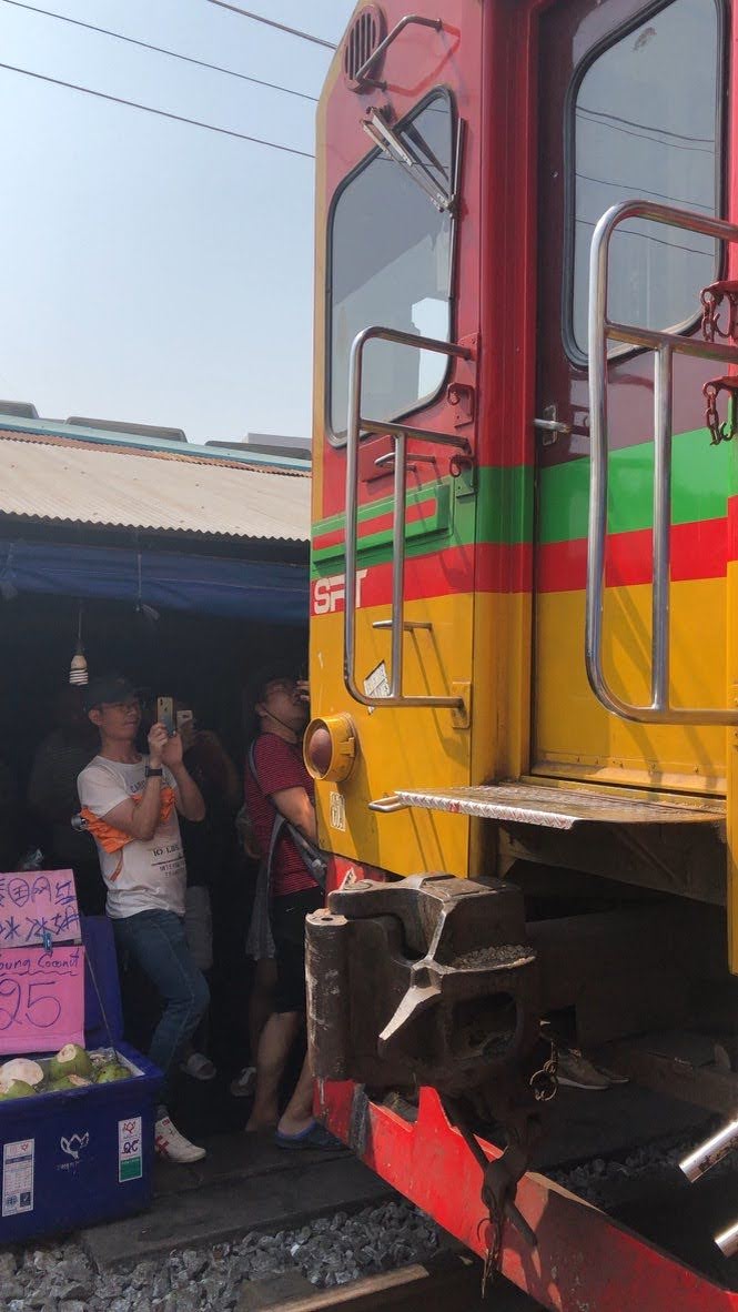 A colorful train passes close to market stalls at Maeklong Railway Market while people watch and take photos.