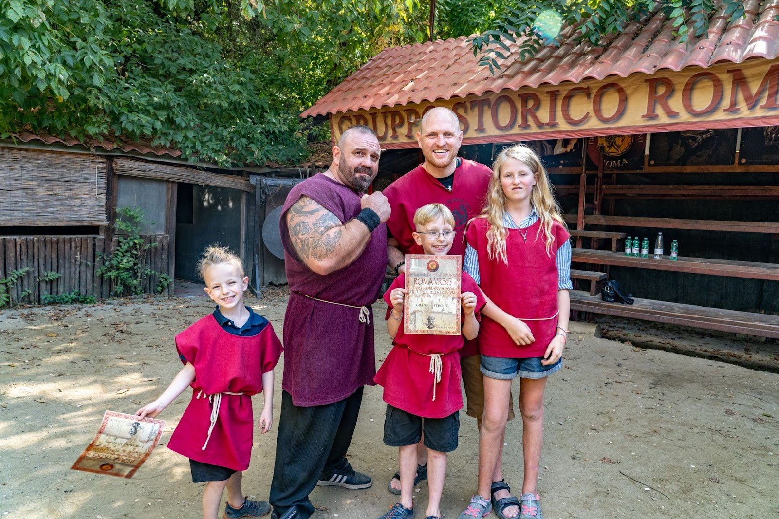 A family in red Roman-style tunics poses with a costumed reenactor in a courtyard.