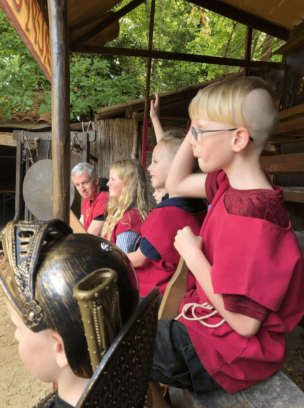 A group in red Roman-style tunics sits together beneath a covered wooden shelter.
