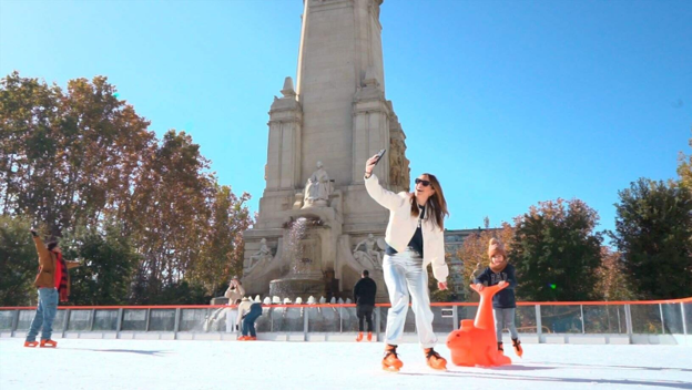 Woman taking a picture on the ice rink, kid to her side, and monument and people behind her