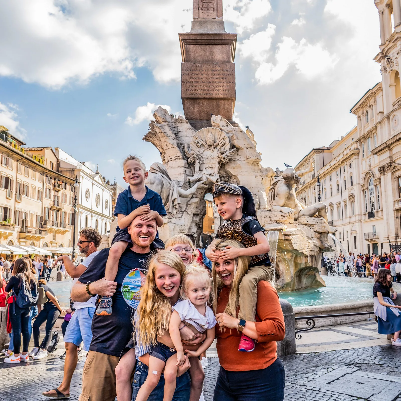 A family poses beside an ornate fountain in a busy Roman piazza.